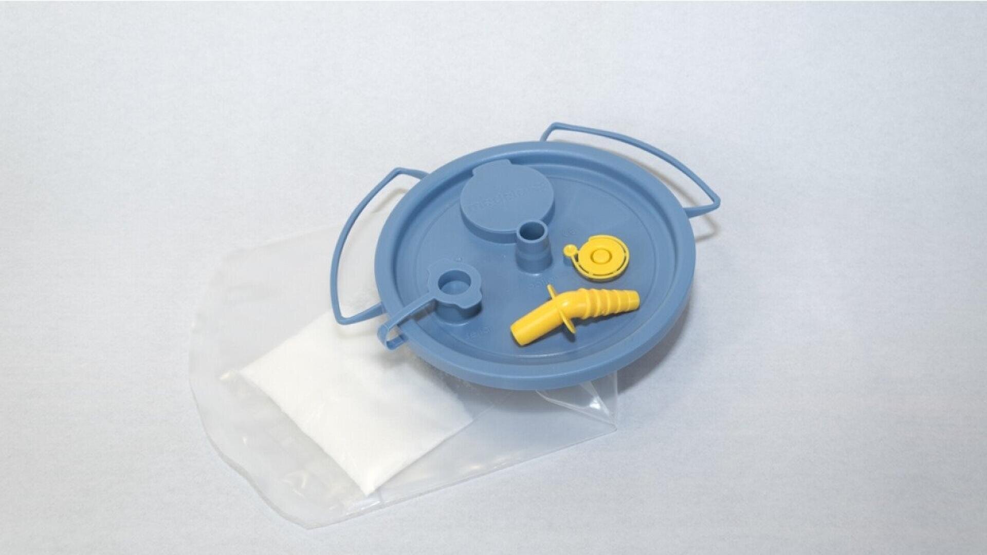 A blue plastic lid with two handles and multiple ports sits on top of a clear bag with a white powdery substance inside. The lid has attached yellow caps and a nozzle. The background is plain and light-colored.