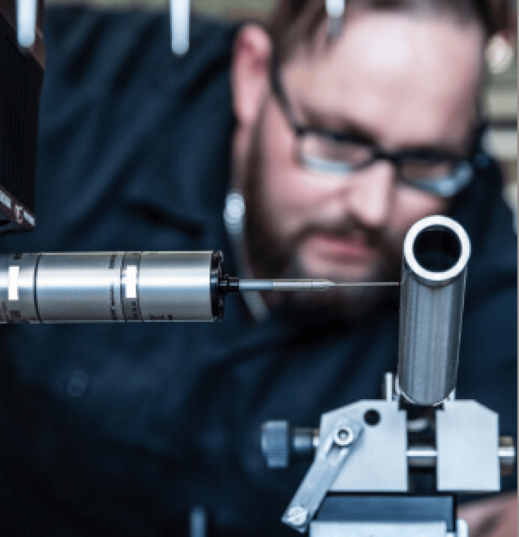 A person wearing glasses closely inspects a metal cylinder held in a clamp, using precise measuring equipment in a workshop or laboratory setting.