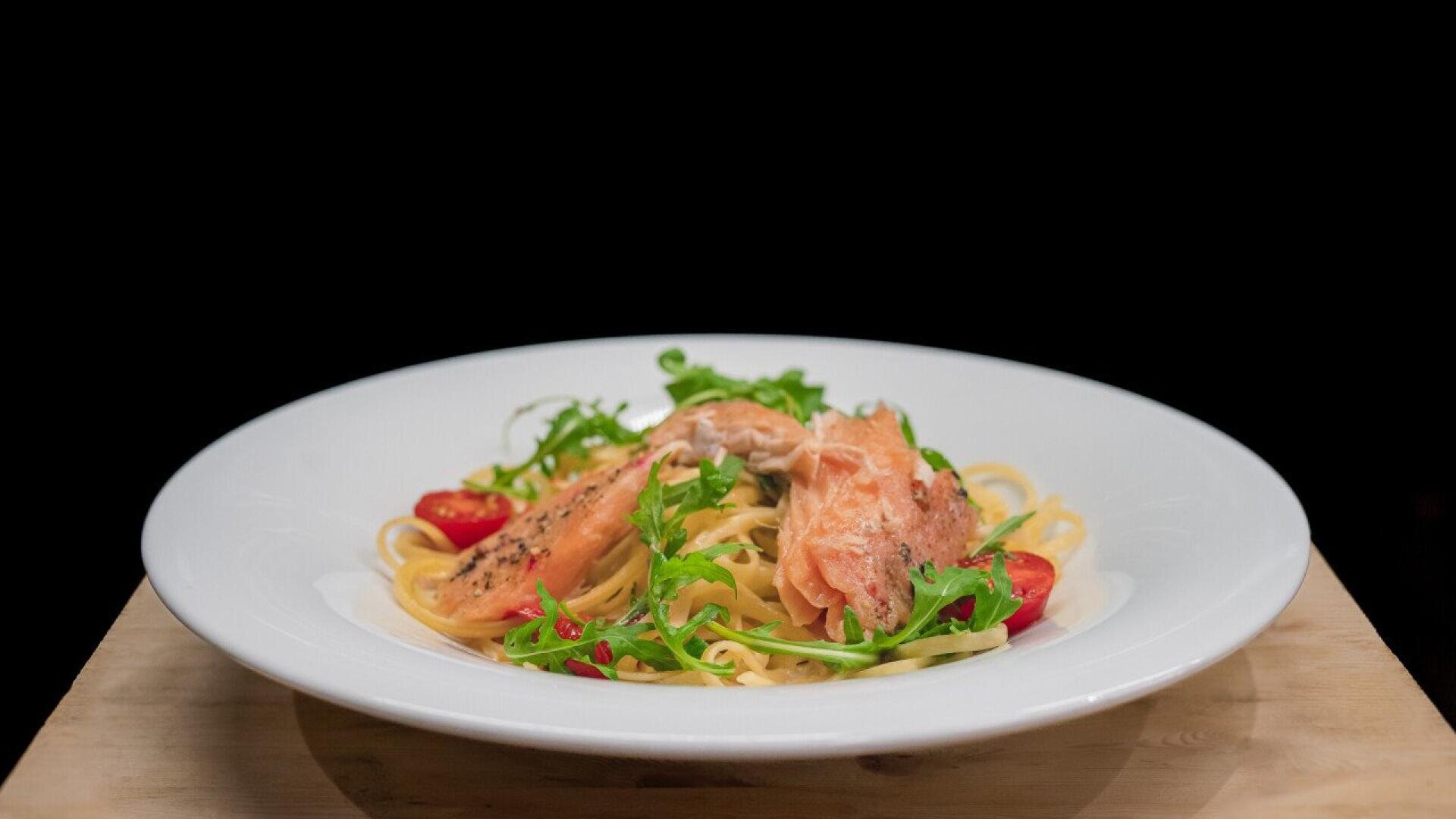 A white plate of pasta topped with pieces of cooked salmon, cherry tomatoes, and fresh arugula, set on a wooden surface against a black background.