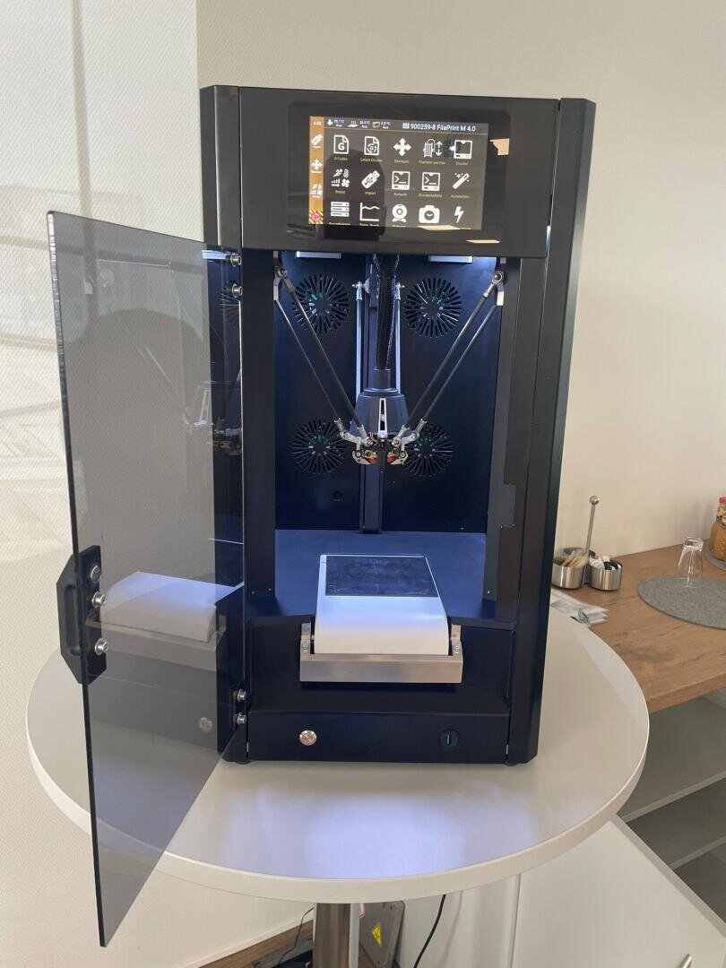 Ein schwarzer 3D-Drucker mit einer Touchscreen-Oberfläche steht auf einem runden weißen Tisch. Die vordere Tür ist geöffnet und gibt den Blick auf den Druckbereich und die internen Komponenten frei. Der Drucker steht in einem modernen, gut beleuchteten Raum.