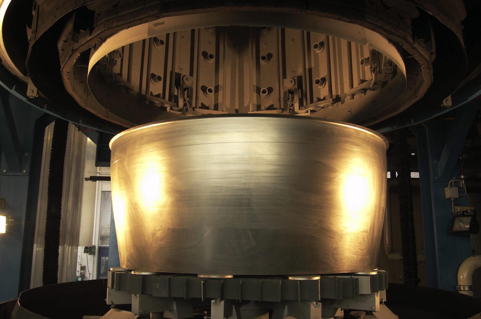 A large cylindrical piece of metal is formed by an industrial machine, surrounded by a circular shape and illuminated by yellowish lights in a factory setting.