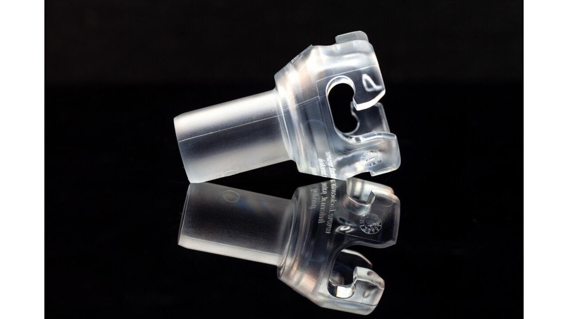 A transparent medical mouthpiece or airway device is shown against a black background, with its reflection visible on the glossy surface below.