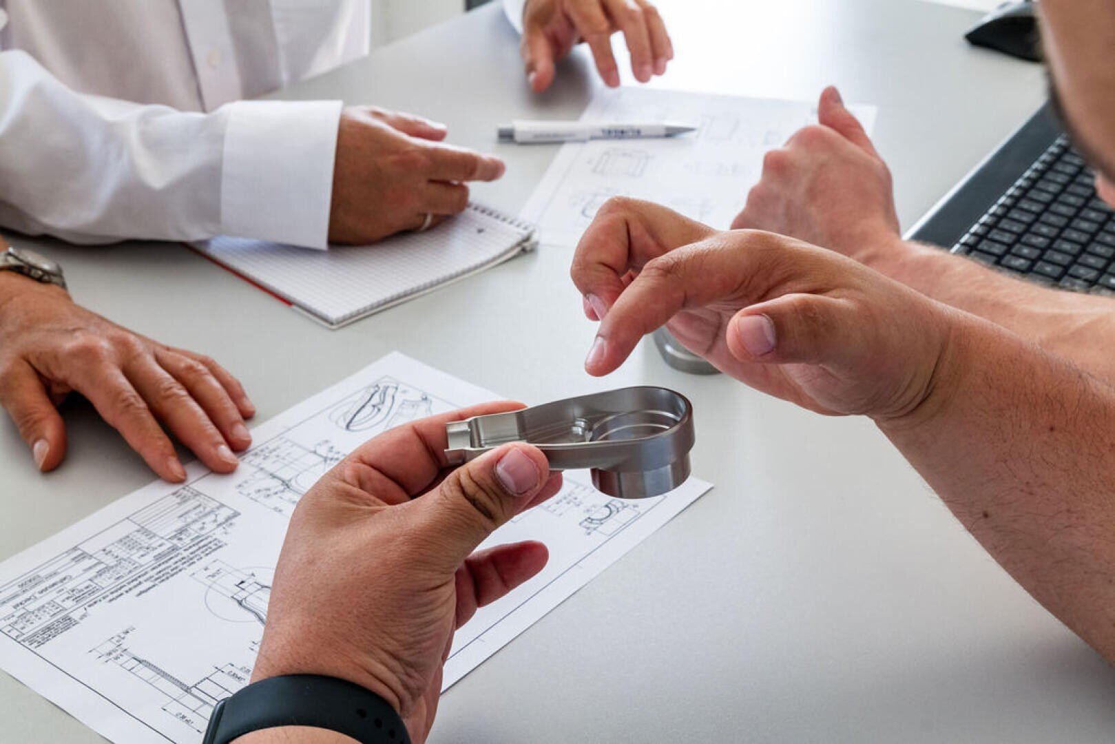 Three people sit at a table and discuss technical drawings while one person holds a metal part in their hand and another examines the features of the part in detail with a gesture.