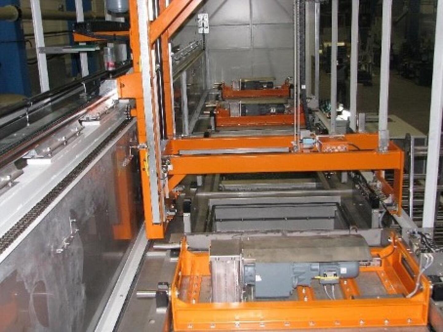 Industrielle Ausrüstung in einer Fabrik, mit orangefarbenen Metallrahmen, Förderbändern und Maschinen, die an einem automatisierten Fertigungs- oder Montageprozess beteiligt sind. Der Bereich ist gut beleuchtet, und verschiedene mechanische Teile sind sichtbar.