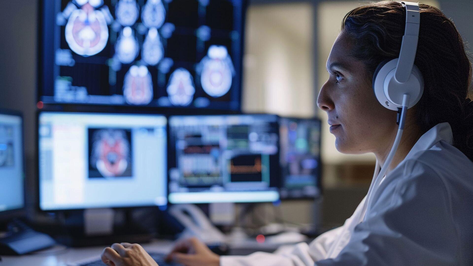 A person in a white lab coat and headphones sits at a desk analyzing brain scans and medical data displayed on several computer monitors in a dimly lit room.