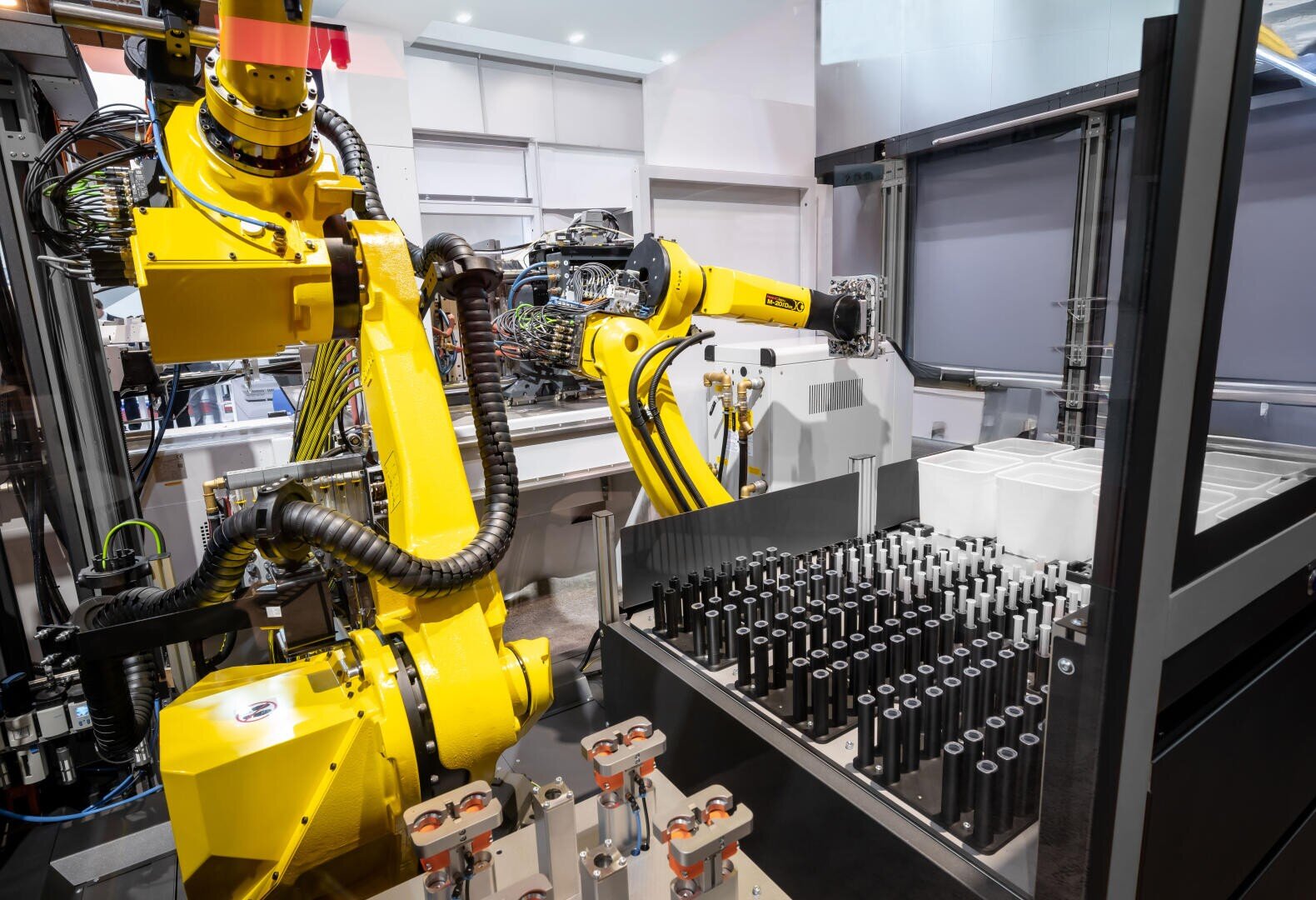 Two large yellow industrial robot arms work in a modern factory and handle cylindrical metal parts in a production line with precision. Various machines and storage areas can be seen nearby.