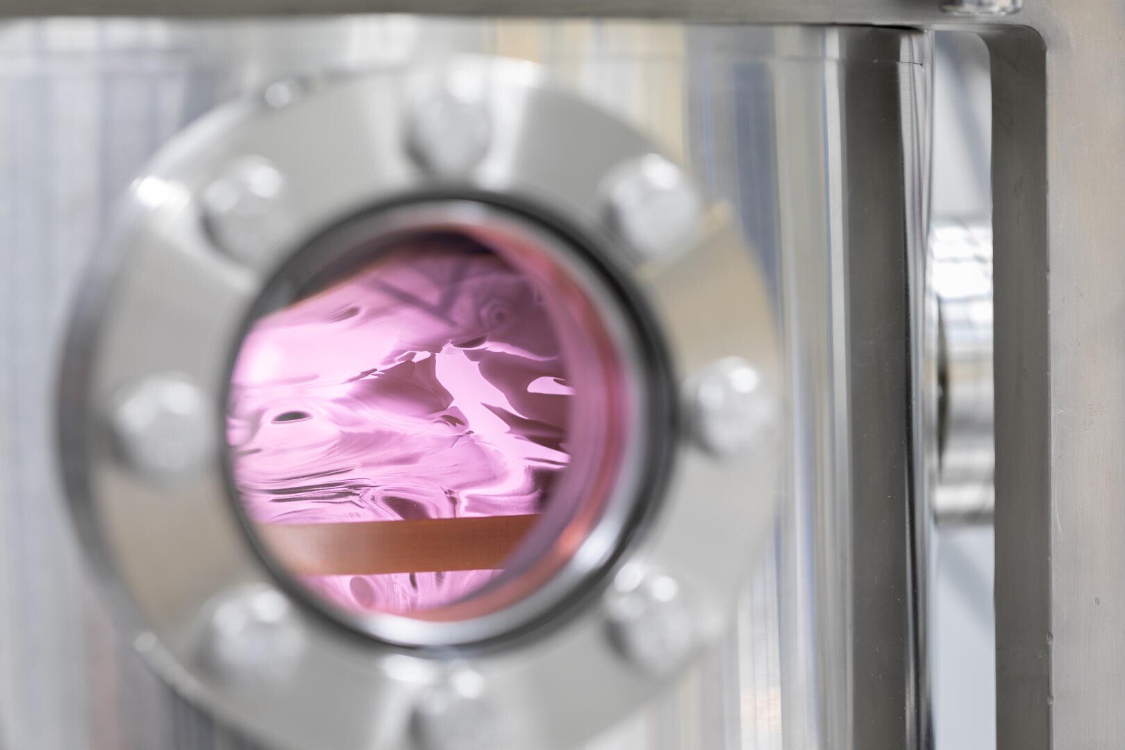 A close-up of a round metal window on a stainless steel chamber revealing a glowing pink plasma or gas inside, with reflections and bolts visible on the metal surface.