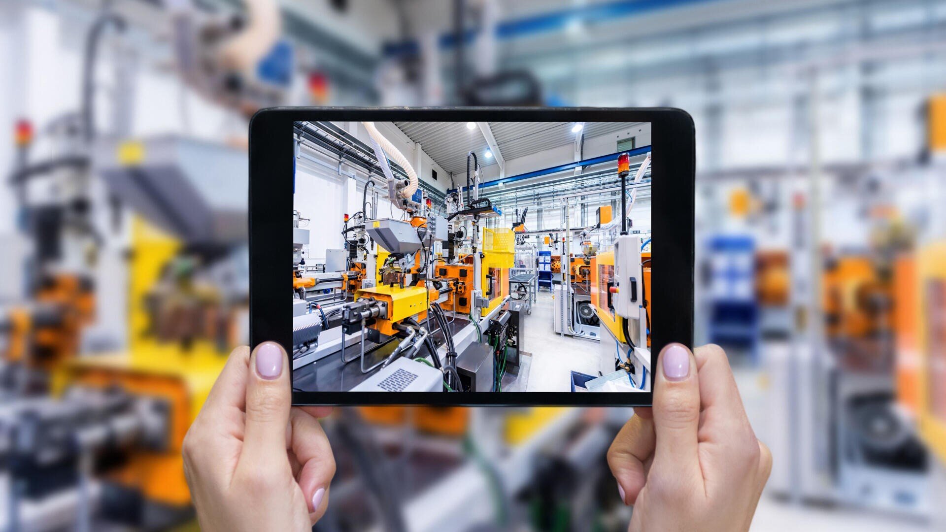 Hände, die ein Tablet halten, das eine Live-Ansicht einer modernen, hell beleuchteten Fabrik oder Industrieanlage mit gelben und orangefarbenen Maschinen zeigt, die sich an den Hintergrundgeräten der Anlage orientieren.