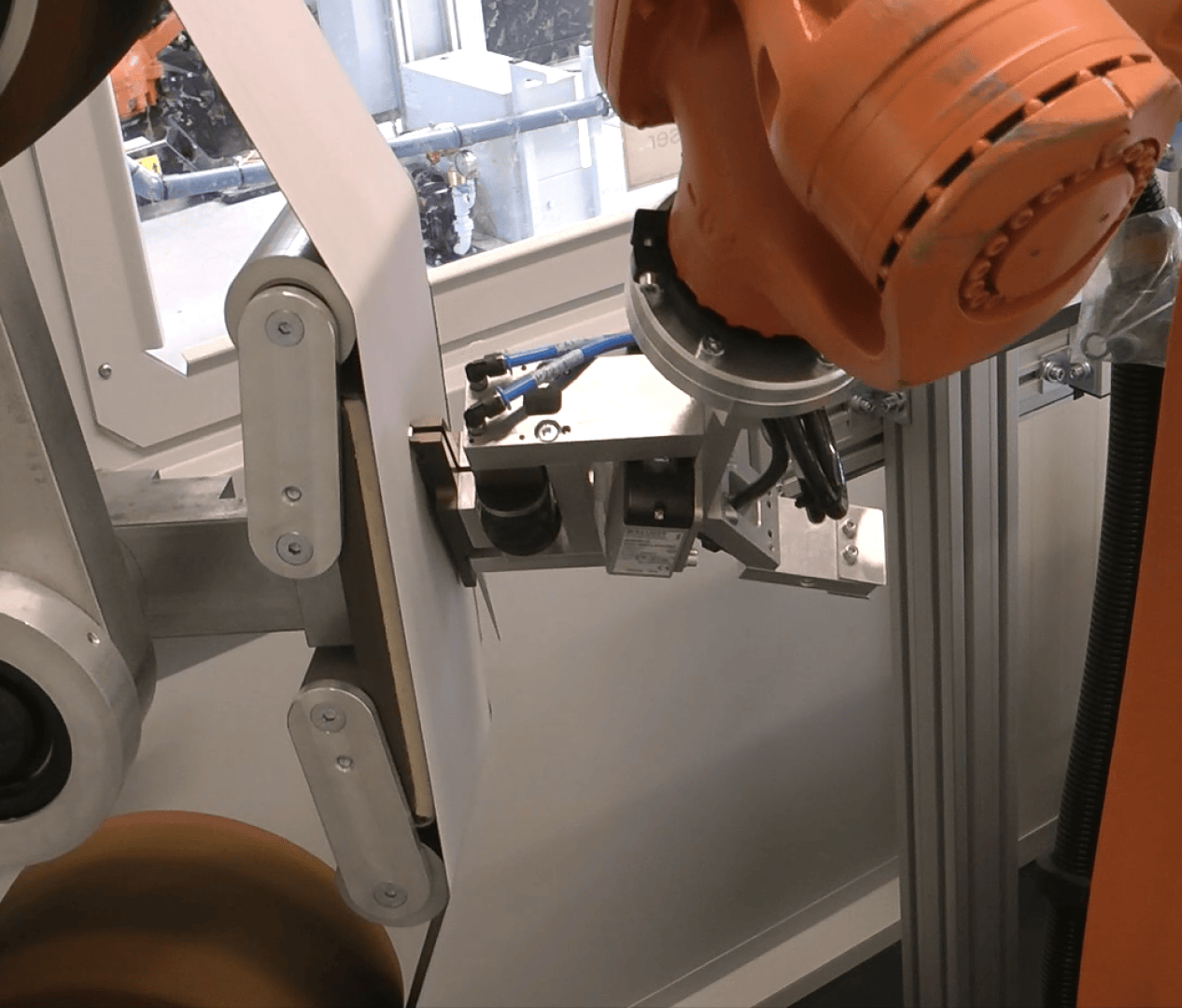 An industrial robotic arm with an orange housing works near a metal fixture in a manufacturing or assembly environment. Machines and a window to another work area can be seen in the background.