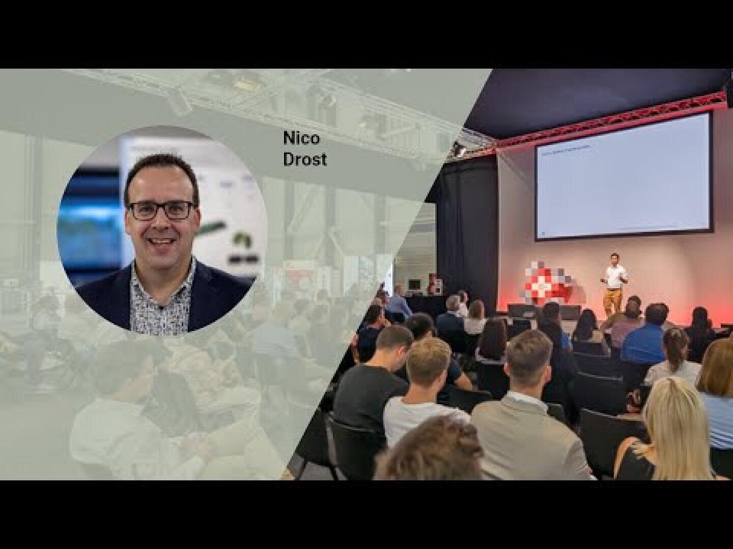 Ein Mann steht auf der Bühne und hält eine Präsentation vor einem sitzenden Publikum in einem modernen Veranstaltungsraum. Ein Overlay zeigt ein Kopfbild eines lächelnden Mannes mit der Bezeichnung Nico Drost auf der linken Seite des Bildes.