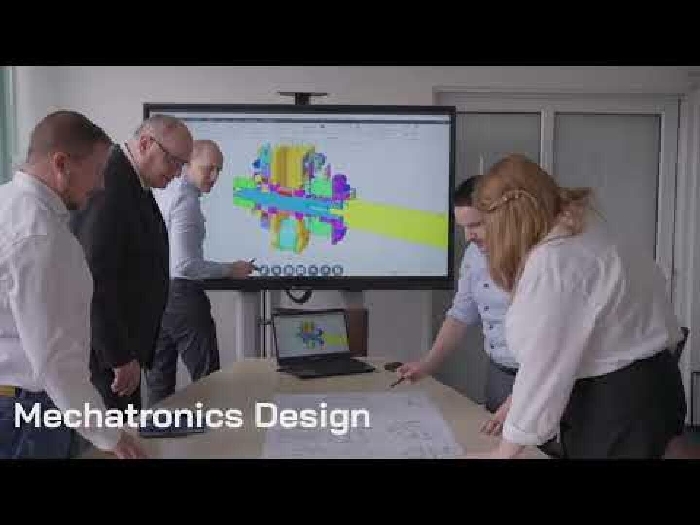 Four people are looking at technical drawings on a table while a screen behind them displays a colorful 3D mechanical model. The text on the picture reads Mechatronics Design.