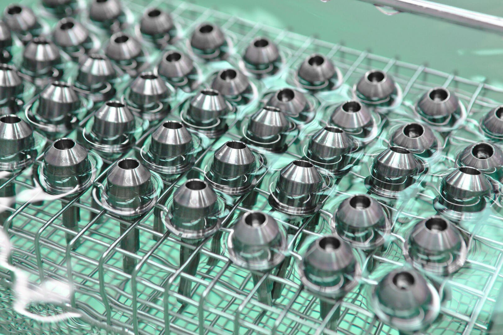 Auf einem Drahtgitter sind Reihen von metallisch glänzenden, kegelförmigen Bauteilen angeordnet, die möglicherweise gereinigt oder bearbeitet werden und von einer grünlichen Flüssigkeit umgeben sind.