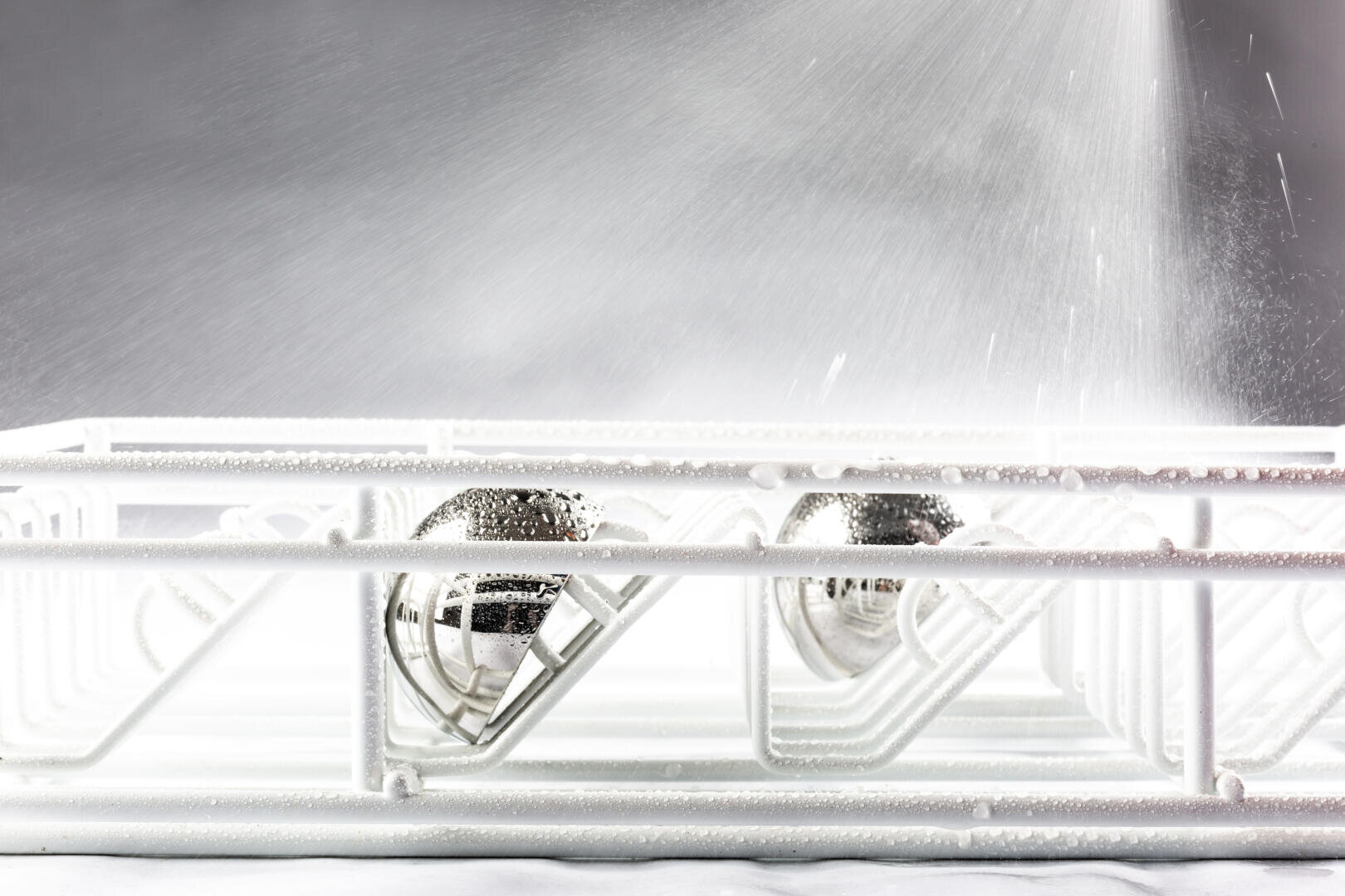 Close-up of a dishwasher rack with two stainless steel balls inside, being sprayed with water from above, creating a misty effect. The scene is brightly lit, highlighting the water droplets and metal surfaces.