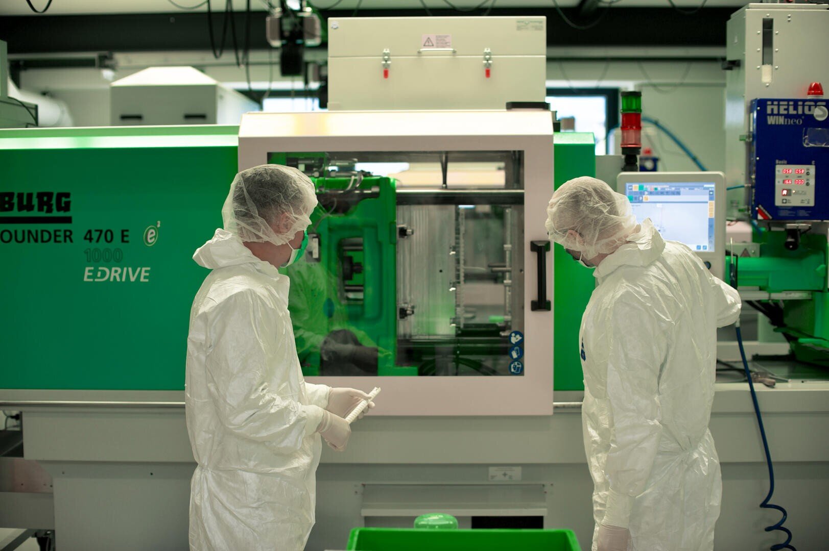 Zwei Personen in weißen Schutzanzügen und Haarnetzen arbeiten an einer grünen Spritzgussmaschine in einem sauberen, modernen Labor oder einer Produktionsstätte. Eine Person hält ein Objekt, während die andere einen Monitor beobachtet.