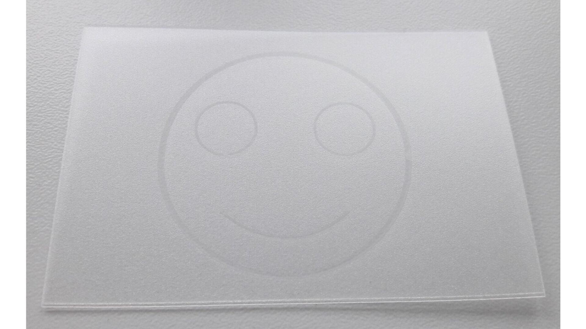 A translucent sheet with a faintly visible smiley face design, placed on a light textured surface. The smiley face consists of two eyes and a curved mouth.