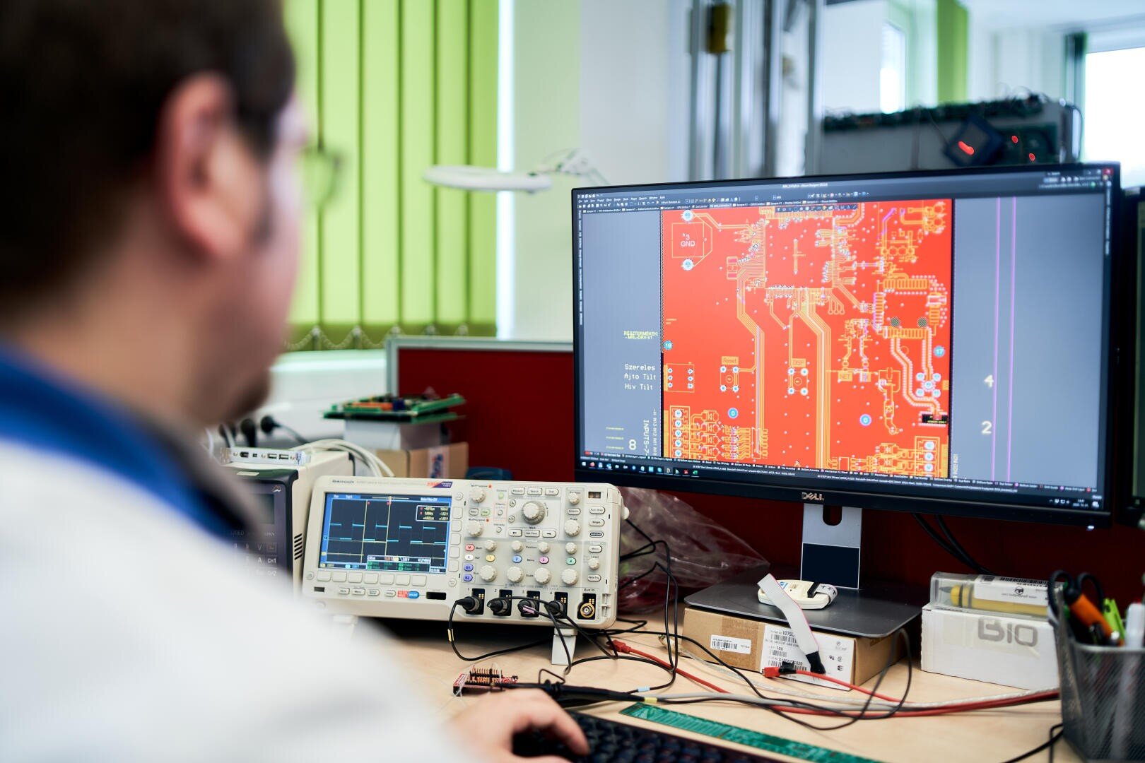 A person sits at a desk working on a computer displaying a detailed circuit board design. Electronic equipment and tools are visible around the workstation in a well-lit office or lab.