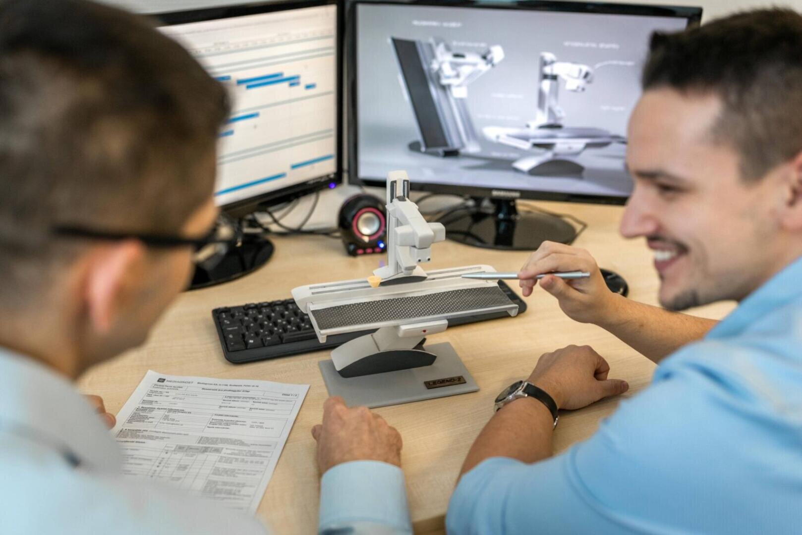 Two men sit at a desk, examining a small mechanical device with a robotic arm. One points at it with tweezers. Computer monitors in the background display technical diagrams and schedules.