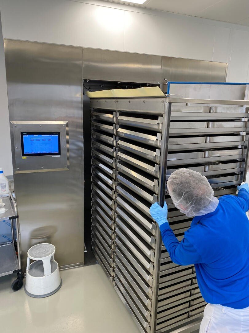 Eine Person in Schutzkleidung und Haarnetz schiebt ein großes Metallgestell mit Tabletts in eine Industriemaschine aus Edelstahl, wahrscheinlich in einem sauberen, sterilen Labor oder einer Lebensmittelverarbeitungsanlage.