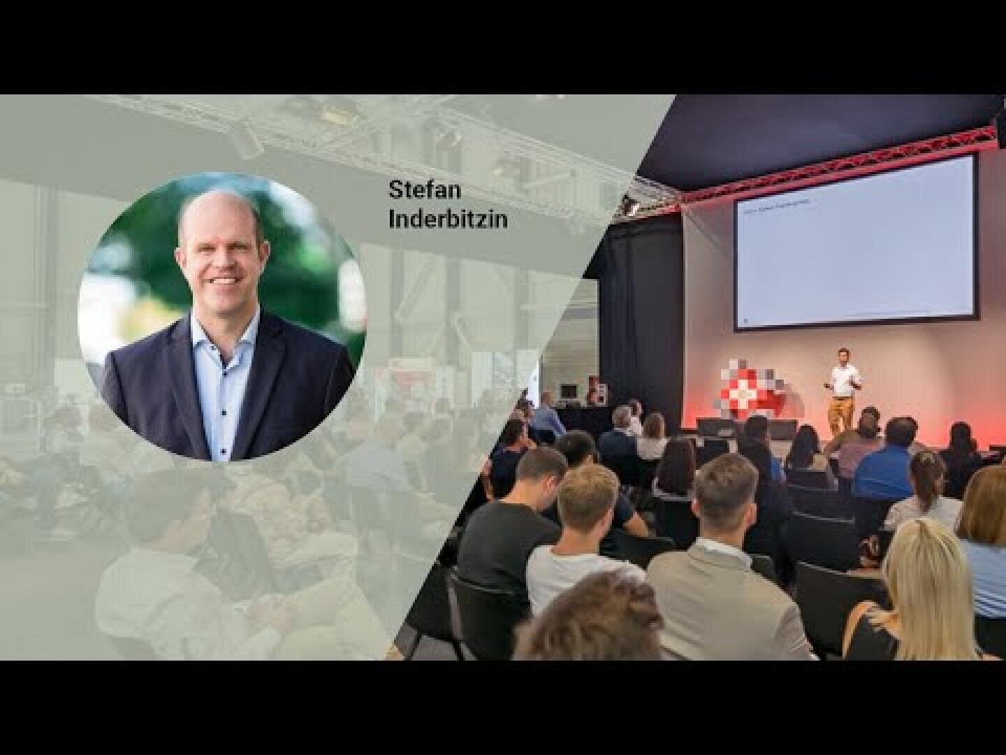 Ein Redner spricht zu einem Publikum in einem Konferenzraum. Auf der linken Seite ist ein kreisförmiges Foto eines lächelnden Mannes im Anzug mit dem Text Stefan Inderbitzin daneben eingefügt.