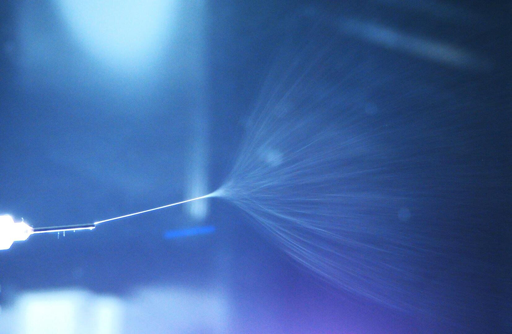 Close-up of a fine nozzle spraying a thin jet of liquid that dissolves into a mist or fine droplets against a blue and violet blurred background.