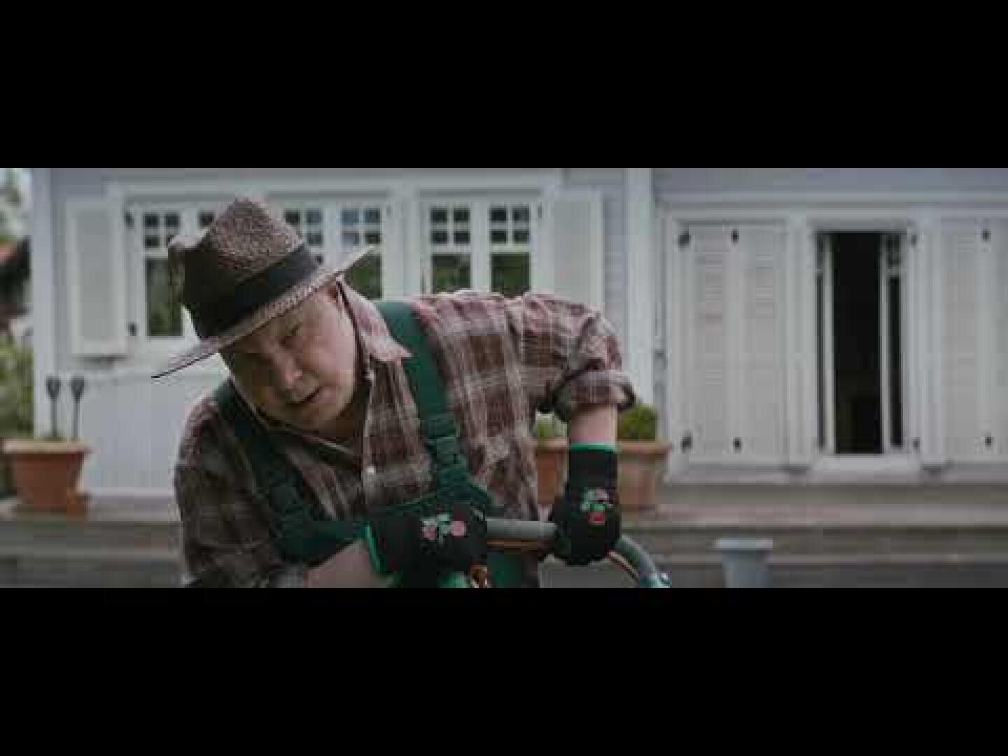 Ein Mann mit Strohhut, kariertem Hemd und grünen Handschuhen arbeitet im Garten vor einem weißen Haus mit großen Fenstern und offenen Fensterläden. Er scheint mit einem Schlauch oder einem Gartengerät zu arbeiten.