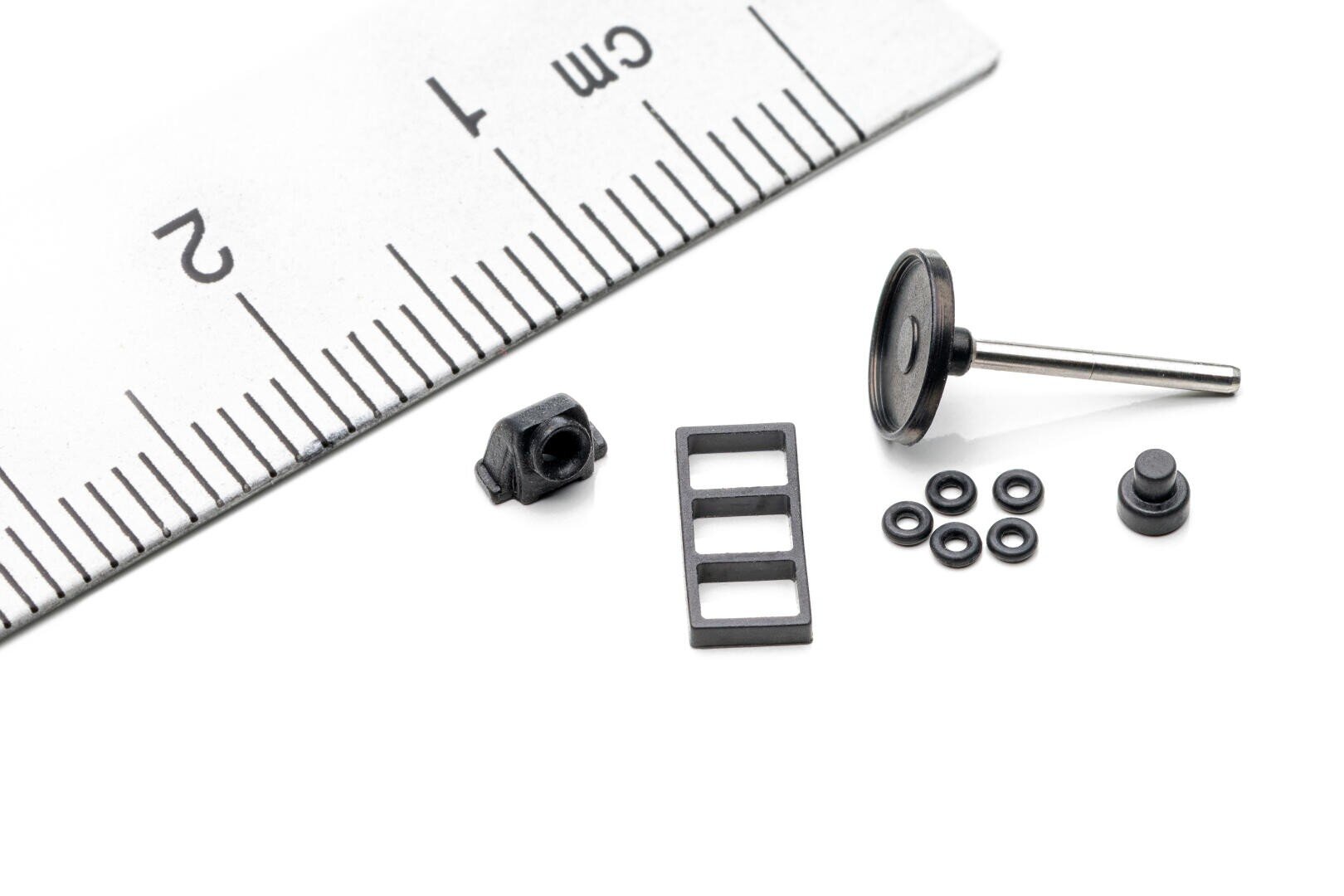 Mehrere kleine schwarze mechanische Teile sind neben einem Metalllineal angeordnet, was ihre geringe Größe unterstreicht. Das Lineal weist Zentimetermarkierungen als Maßstab auf. Der Hintergrund ist weiß.