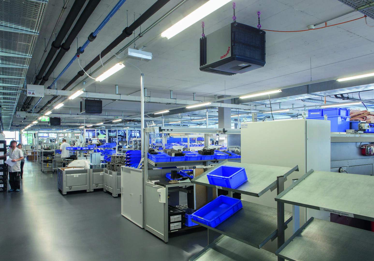 A spacious, modern factory interior with workstations, machines, blue storage boxes and ceiling lighting. Two people in white coats work in the background between organized equipment and shelves.