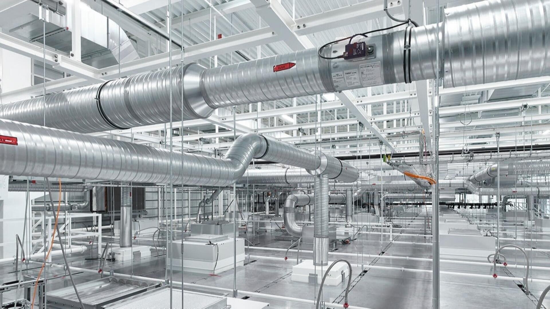 Eine moderne Industrieanlage mit freiliegenden silbernen Luftkanälen, Rohren und Lüftungssystemen, die von der Decke hängen, umgeben von weißen Stahlträgern und heller Beleuchtung.