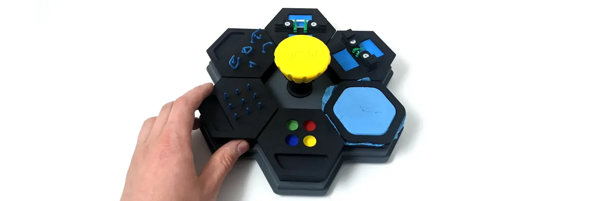 A hand reaches for a black hexagonal controller with a large yellow knob in the center, colorful buttons, and blue tactile elements on a white background.