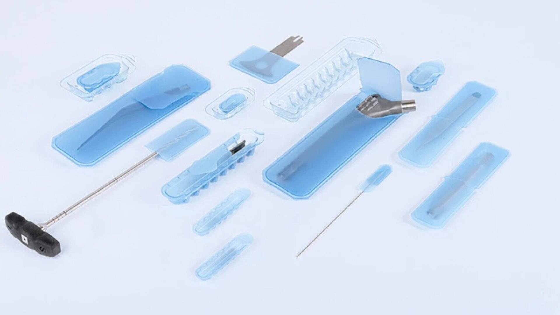 A variety of medical tools and surgical instruments in blue sterile packaging are arranged on a white surface. The items include needles, forceps, and other precision tools commonly used in medical procedures.