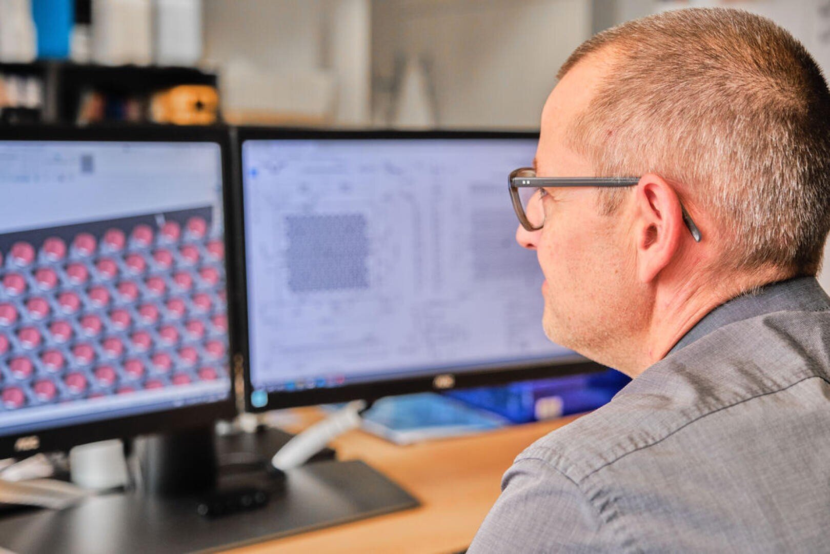 A person wearing glasses sits at a desk and works on two computer monitors displaying technical and 3D modeling designs in an office environment.