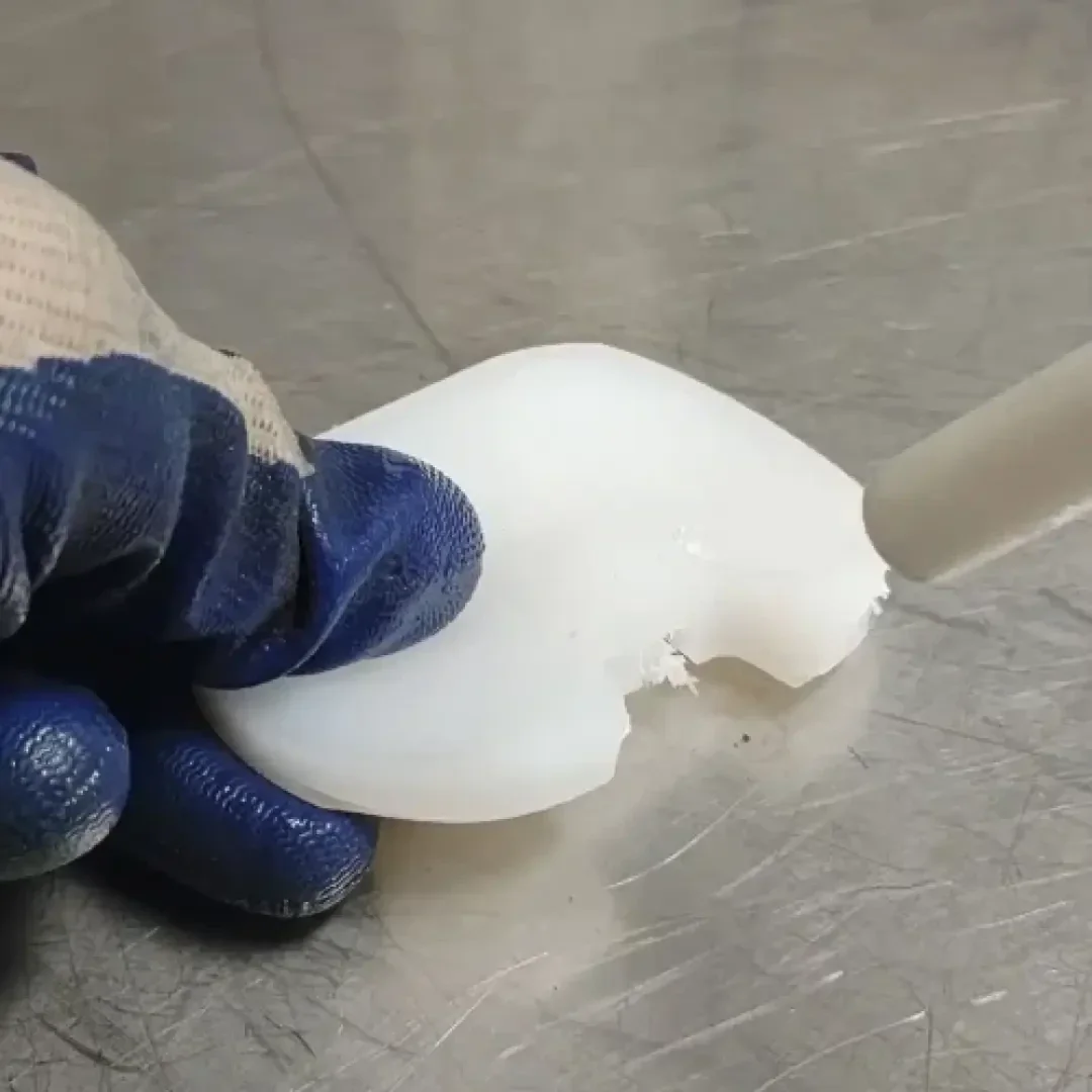 A gloved hand holds a white, translucent, rectangular object on a metal surface while a tool cuts off a piece of its edge.