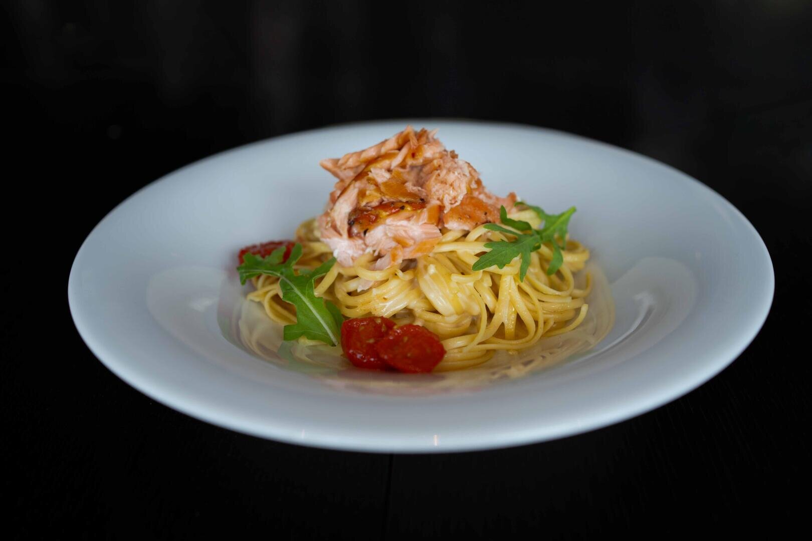 Ein weißer Teller mit Spaghetti, belegt mit gekochten Lachsstücken, Kirschtomaten und Rucolablättern, serviert auf einer dunklen Unterlage.