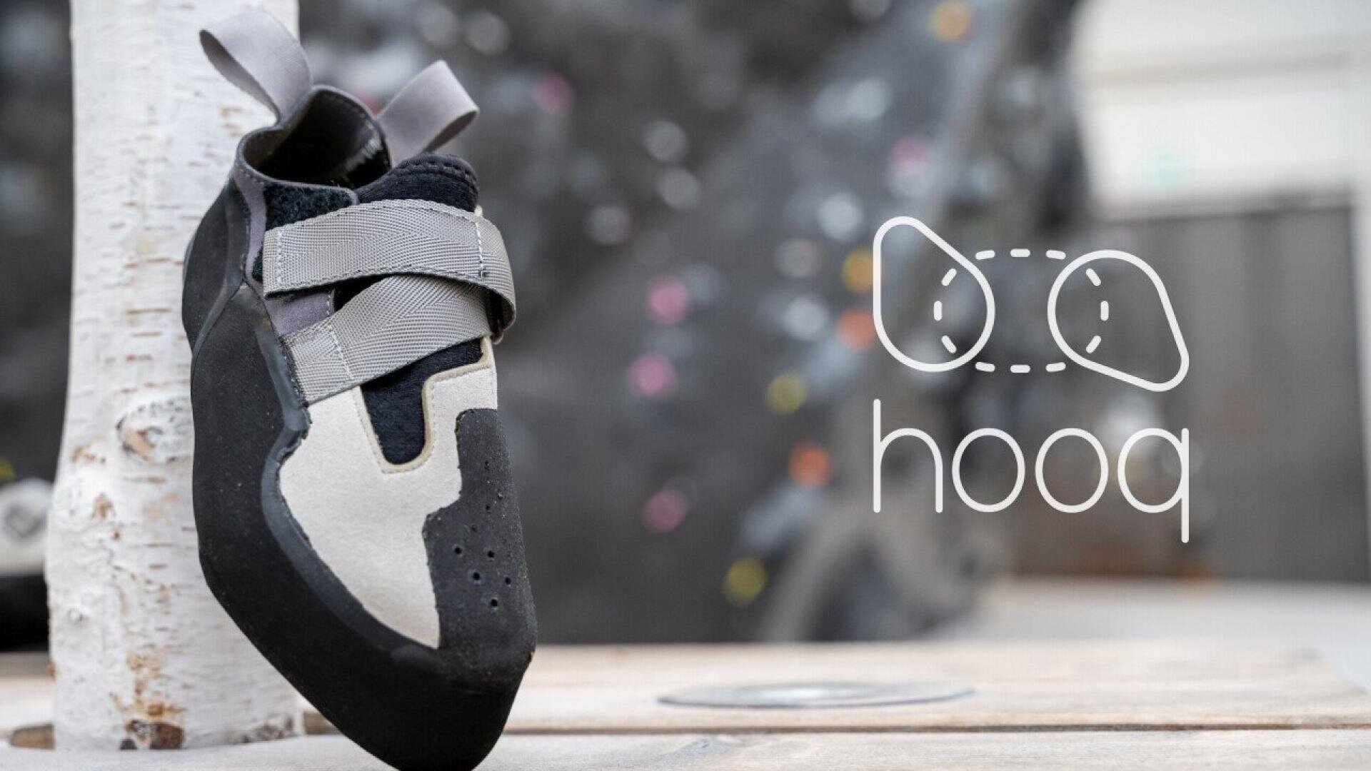 A gray and black climbing shoe with two Velcro straps rests against a white birch log. In the background, the word hooq and a stylized logo are visible, along with an indoor climbing wall.