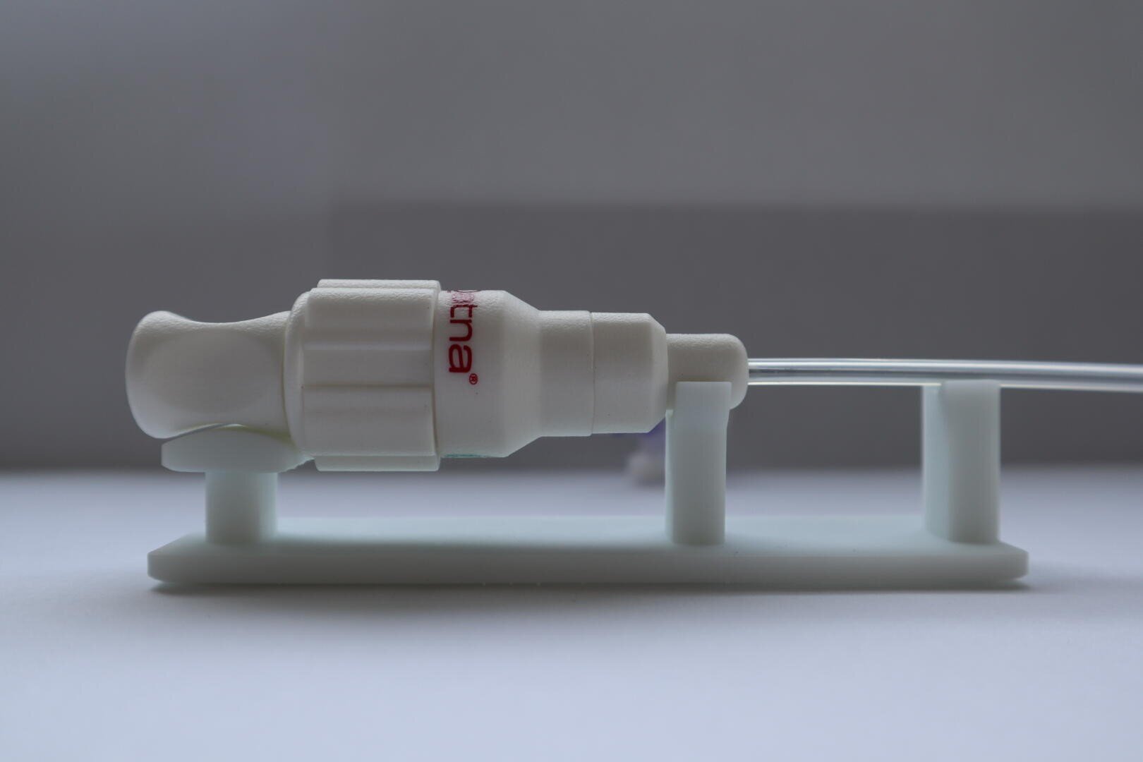 Close-up of a medical device with a white plastic connector attached to a transparent tube and resting on a white stand. The connector is partially labeled in red and photographed against a plain grey background.