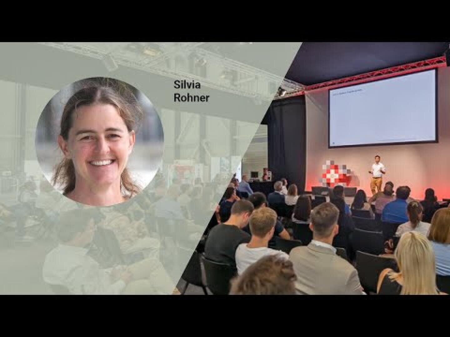 Eine Frau namens Silvia Rohner ist lächelnd in einem eingeschnittenen Kreis auf der linken Seite zu sehen. Auf der rechten Seite spricht ein Mann auf der Bühne zu einem Publikum in einem modernen Konferenzraum mit einem großen Bildschirm hinter ihm.
