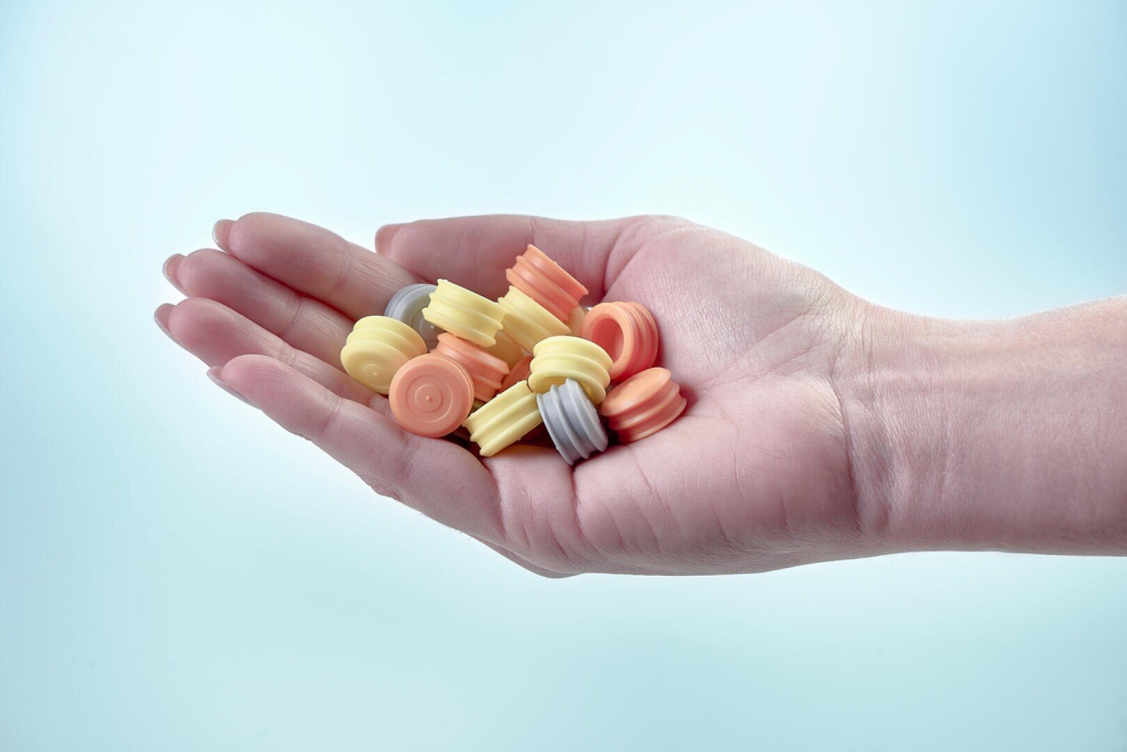 One hand holds several colorful, round, ribbed sweets in shades of yellow, orange, grey and peach against a pale blue background.