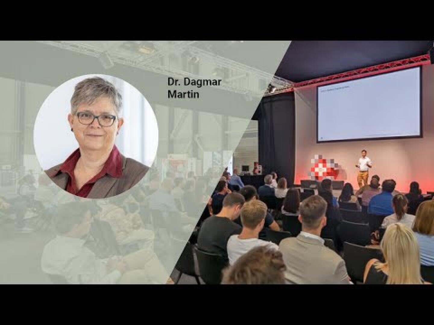 Ein großes Publikum sitzt vor einer Bühne, auf der ein Redner neben einer Leinwand spricht. Auf der linken Seite ist ein rundes Foto von Dr. Dagmar Martin eingefügt, neben dem ihr Name steht.