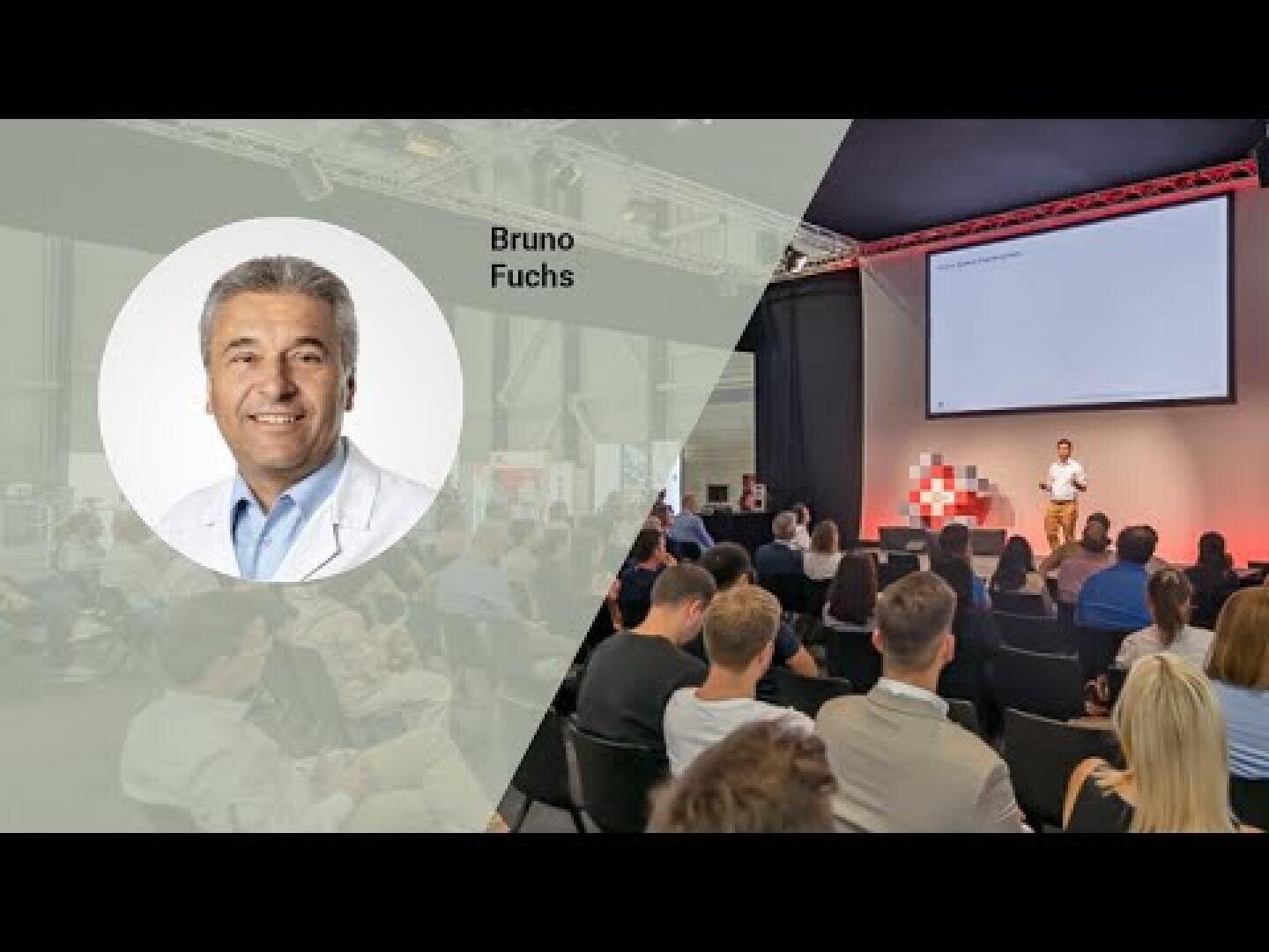 Ein Mann steht auf der Bühne und hält einen Vortrag vor dem Publikum; links daneben ist das Porträt eines lächelnden Mannes in einem weißen Kittel mit der Aufschrift Bruno Fuchs zu sehen.