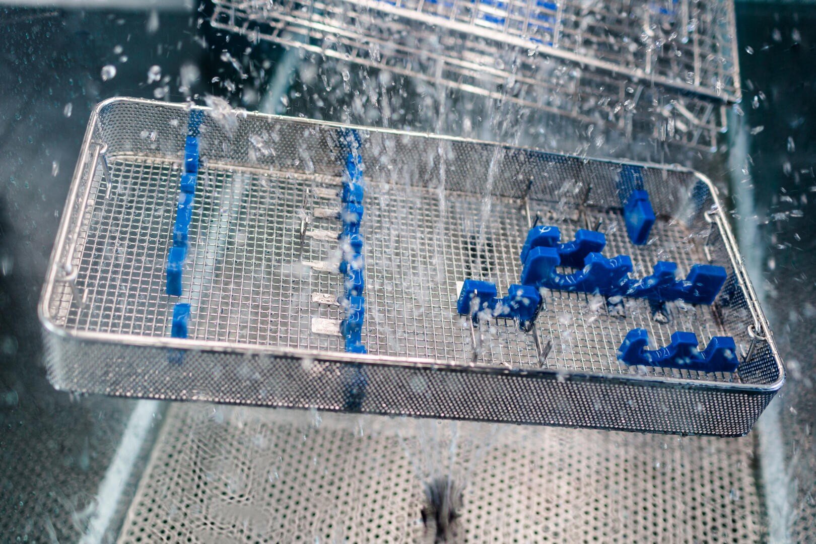 A metal tray with blue plastic holders is being sprayed with water, likely in a sterilization or cleaning process, inside a metal container or machine.
