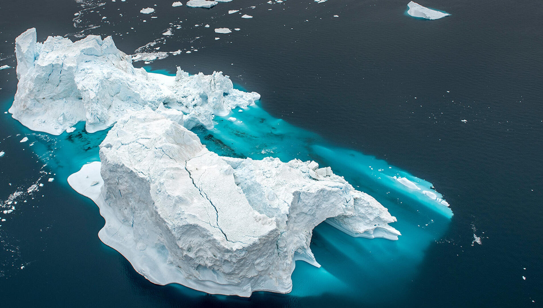 A large white iceberg floats in dark blue water, with bright turquoise patches around its base, indicating shallow, submerged ice. Smaller pieces of ice float nearby under a cloudy sky.