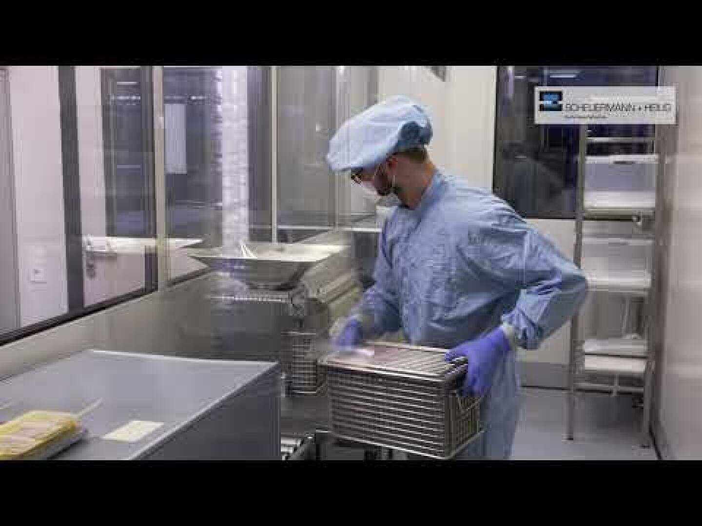 A person in a blue sterile suit, mask, and gloves handles a metal container inside a cleanroom laboratory with stainless steel equipment and glass walls.