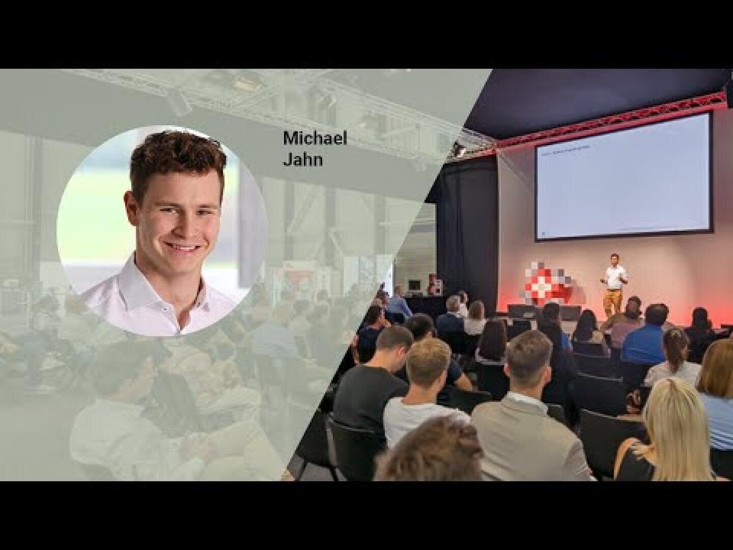 Ein Mann, der als Michael Jahn identifiziert wurde, wird gezeigt, wie er auf der Bühne zu einem Publikum in einem Konferenzraum spricht. Auf der linken Seite befindet sich ein kreisförmiger Einschub mit seinem Porträt und seinem Namen.