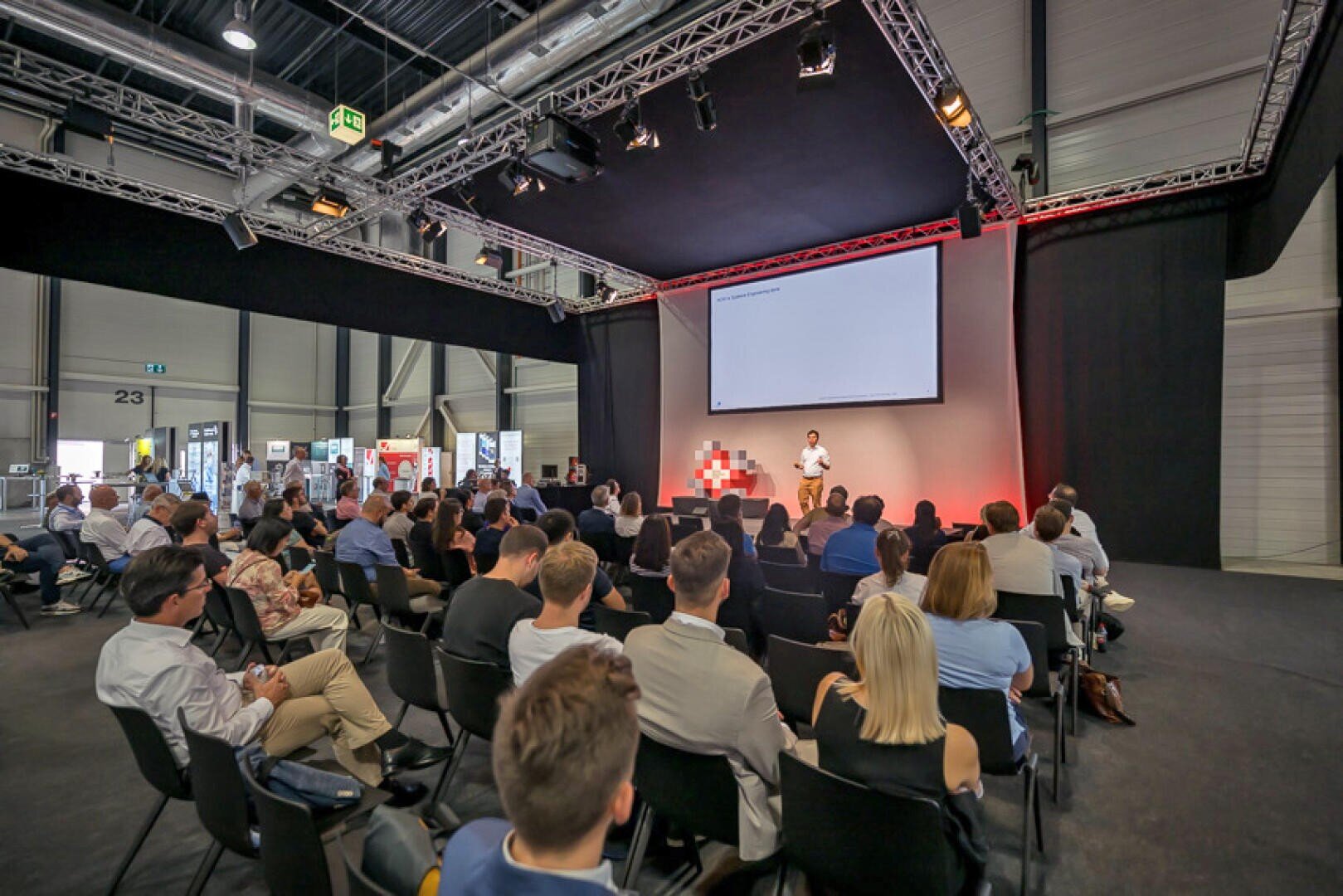 Auf einer Konferenz oder einem Seminar spricht ein Redner auf der Bühne zu einem in Reihen sitzenden Publikum, hinter dem eine große Projektionsfläche und im Hintergrund Ausstellungsstände zu sehen sind.