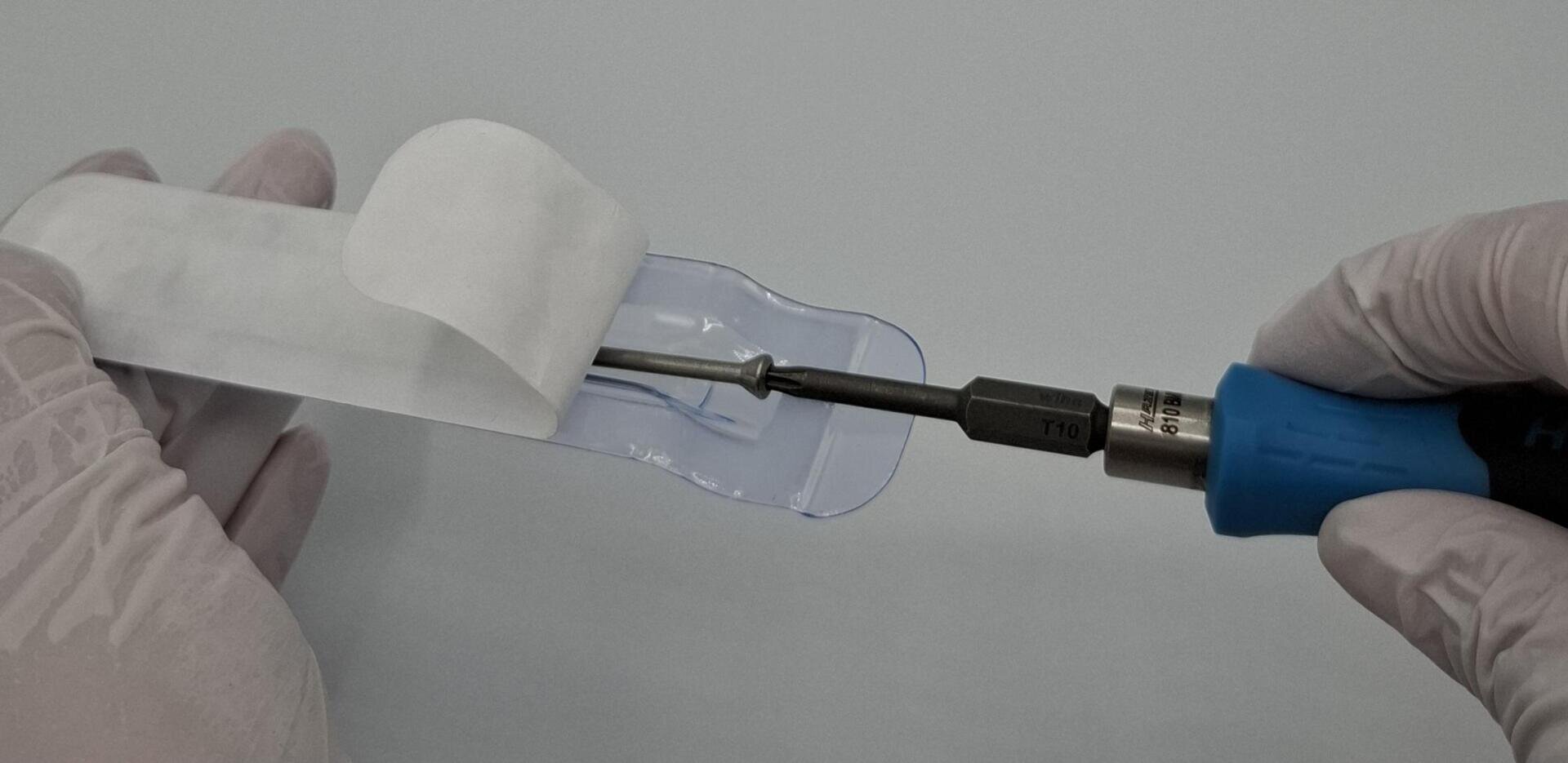 A gloved hand holds a screwdriver with a blue handle and inserts a metal tool into a transparent plastic holder that is partially covered with white paper. The background is monochrome light gray.