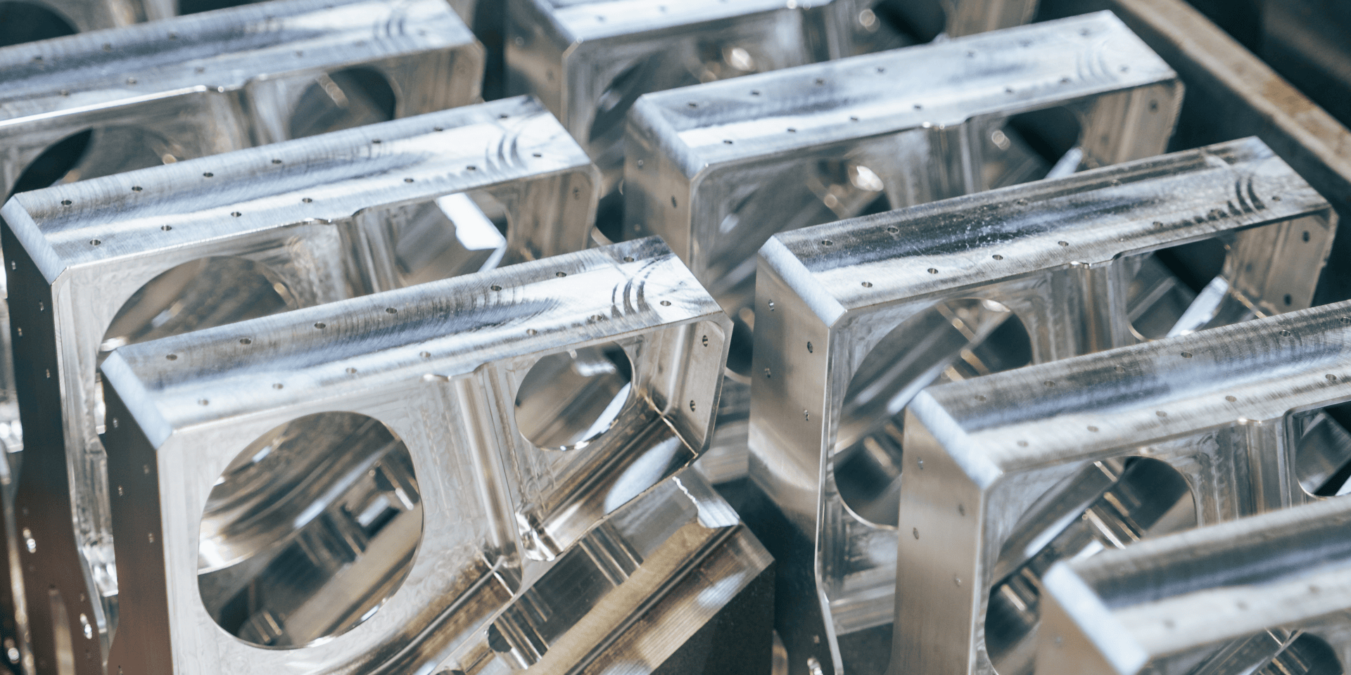 Rows of shiny, rectangular, CNC-milled metal parts with precise holes and edges that are closely aligned and reflect the light on their polished surfaces.