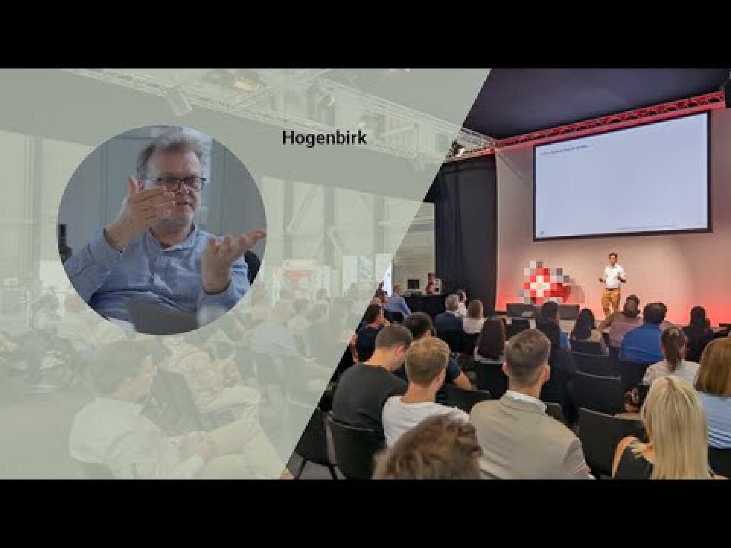 Ein Mann hält einen Vortrag vor einem sitzenden Publikum in einem Konferenzraum. Ein Einsatz zeigt einen anderen Mann, der die Gebärdensprache verwendet, mit der Aufschrift Hogenbirk. Auf der Bühne sind ein Bildschirm und rot-weiße Blöcke zu sehen.