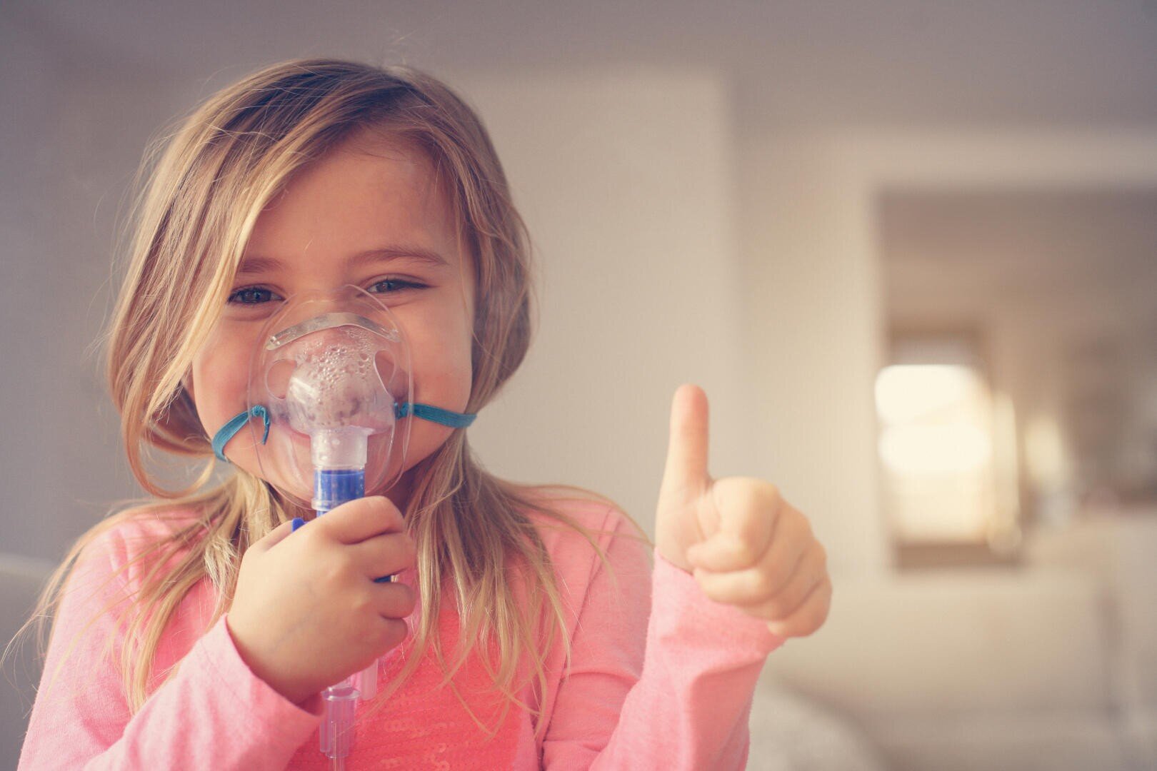 A young girl with long blonde hair wears an inhalation mask and points her thumb upwards with her other hand. She is smiling and wearing a pink shirt. She is sitting inside in a dimly lit room.