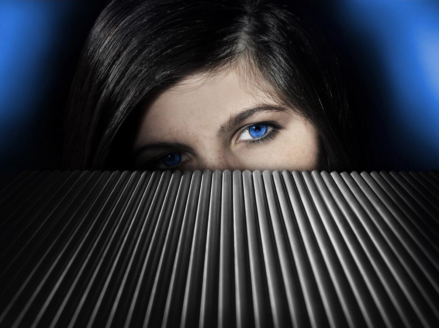 A person with striking blue eyes and dark hair stares ahead, her face partially hidden behind a metallic, ribbed surface with a dramatic blue and black background.