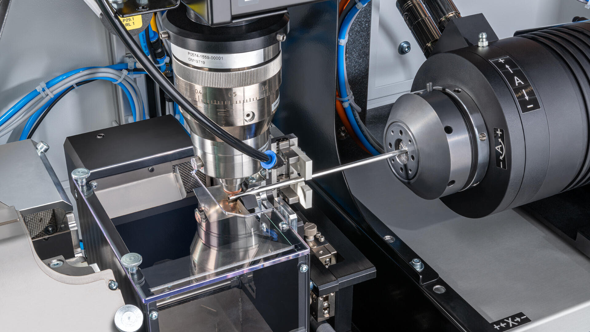 Nahaufnahme einer CNC-Maschine in Betrieb, die eine Metallstange zeigt, die von rotierenden Schneidwerkzeugen präzise bearbeitet wird, umgeben von Drähten und verschiedenen Maschinenkomponenten.